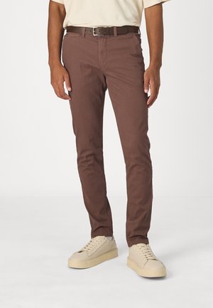 Mężczyzna w wąskich brązowych spodniach, ciemnobrązowym pasku i beżowych trampkach, stojący na jednolitym białym tle.