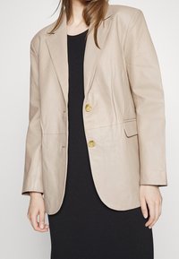 Béžová kožená blazer s jednopropínacím designem, dvěma zlatými knoflíky a texturovaným černým tričkem. Hladký povrch, přizpůsobený střih.