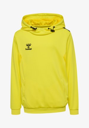 Niño de pie vistiendo una sudadera deportiva con capucha de color amarillo brillante y pantalones cortos, calcetines negros y zapatos deportivos negros sobre un fondo gris liso.