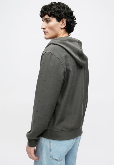 Χακί φούτερ με υφαντό μοτίβο και κουκούλα με κορδόνια, συνδυασμένο με ανοιχτό μπλε παντελόνι τζιν. Απλή, άκοπη κατασκευή.