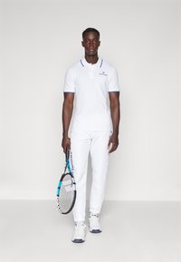 Maglietta polo bianca con dettagli blu e logo sul petto, abbinata a pantaloni bianchi e sneakers bianchi. Il modello tiene una racchetta da tennis.