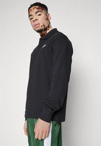 Camisa polo de manga longa preta em mistura de algodão, com gola dobrada e um pequeno logotipo branco no lado esquerdo do peito, combinada com calções verdes.
