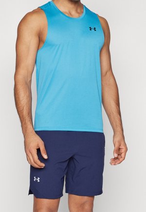 Mężczyzna w niebieskim sportowym topie bez rękawów i granatowych spodenkach Under Armour, stojący z rękami swobodnie opuszczonymi wzdłuż ciała.