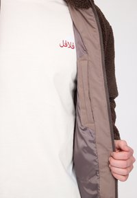 Brown fleece jacket with a zipper, featuring a smooth inner lining and a white shirt underneath with red Arabic text.