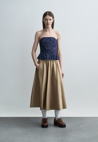 Kvinne som står iført en mørkeblå stroppeløs polkadottopp, beige midikjole, hvite sokker og brune platåsko mot en ensfarget bakgrunn.