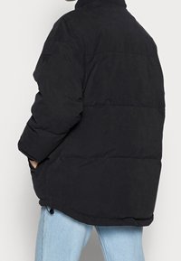 Persona vestida con una chaqueta acolchada negra, con las manos en los bolsillos, combinada con jeans celestes, de pie contra un fondo liso.
