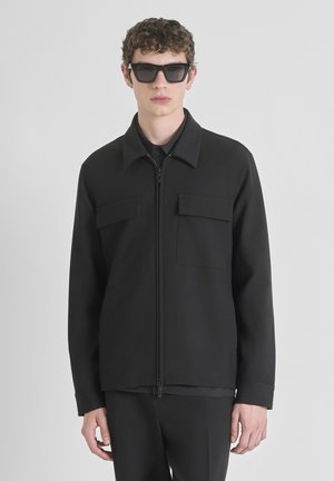 Veste zippée noire en tissu lisse, avec un col pointu et deux poches poitrine. Portée avec un pantalon coordonné et des lunettes de soleil.