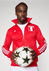 Punane lukuga jakir, millel on valged Adidas triibud, ning Peruu lipu plaastriga. Hoides valget jalgpalli mustade tähtedega.