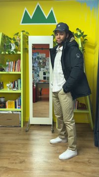 Fekete pufi dzseki, kockás ing, khaki nadrág, fehér tornacipő. A háttérben zöld könyvespolc, tükör és sárga fal látható hegy mintával.