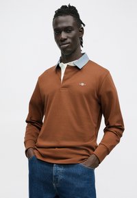 Bruine polo met lange mouwen, een lichte denimkraag en een witte knoopsluiting; heeft een klein geborduurd logo op de borst.