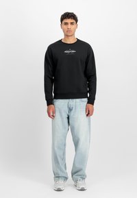 Melnā sporta krekls ar apaļu kakla izgriezumu un logo uz krūtīm, kombinēts ar gaiši zilām brīvām džinsu biksēm un pelēkiem sporta apaviem. Vienkrāsains, ikdienišķs dizains.