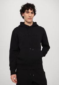 Hanorac negru cu glugă dintr-un material moale, având un buzunar frontal tip kangaroo, șnururi de strangulare și un logoembossat al mărcii pe piept.
