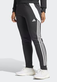 Pantalons de sport noirs avec des panneaux blancs, présentant trois bandes blanches sur les côtés et un logo Adidas. Fabriqués en tissu extensible.