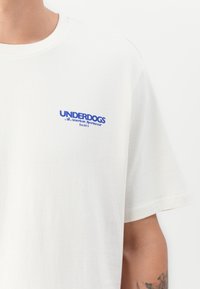 Hvit t-skjorte med blå tekst "UNDERDOGS All American Sportswear Est. 2013" på venstre bryst, båret av en person med tatovert arm.