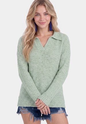 Donna bionda che indossa un maglione a coste verde chiaro e pantaloncini blu navy consumati, sorridente con le mani giunte davanti.