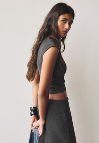 Haut en tricot côtelé gris et jupe assortie. La tenue présente un design court avec une silhouette ajustée. Le mannequin tient un sac à main bleu avec des clous décoratifs.
