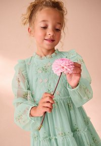 Una giovane ragazza che indossa un vestito a fiori verde menta tiene delicatamente e guarda un fiore rosa su un lungo gambo, contro uno sfondo beige morbido.