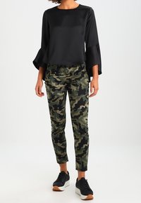 Svart satin topp med utställda ärmar, matchad med åtsittande kamouflagebyxor. Modellen bär svarta sneakers med vita sulor och en ljusbrun accent.