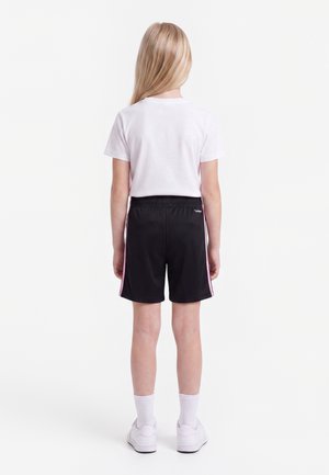 Blond kind staat met de rug naar de camera, draagt een wit T-shirt, zwarte korte broek met roze strepen, witte sokken en witte sneakers tegen een effen achtergrond.