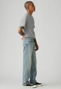 Hombre con tatuajes que lleva una camiseta gris, vaqueros claros y zapatillas negras, de pie de perfil sobre un fondo liso.