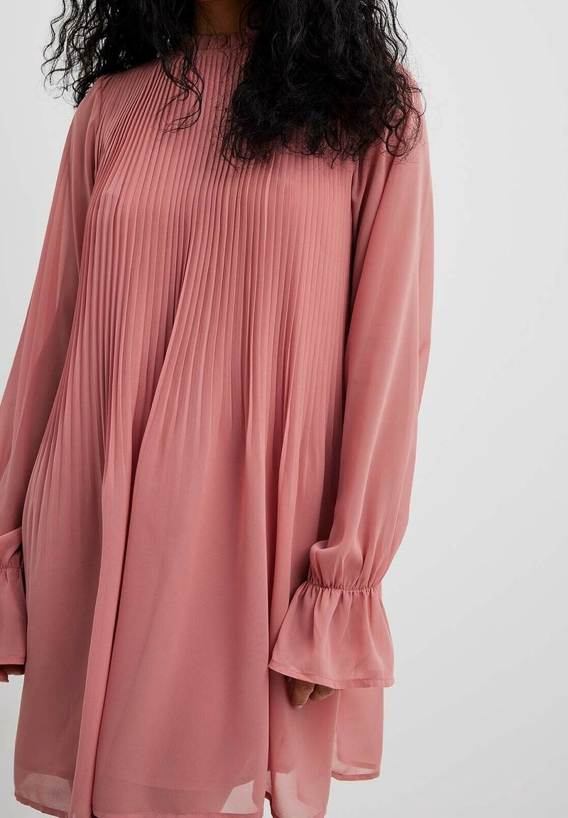 Femme portant une robe ample rose poussière à manches longues avec des plis verticaux sur le haut devant et des poignets évasés.