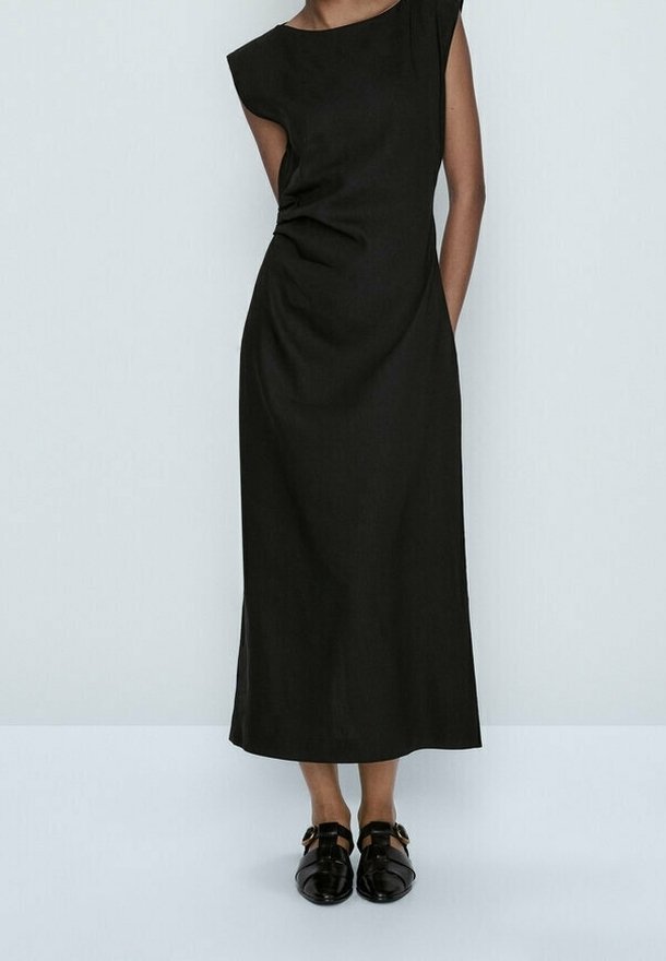 Femme portant une robe midi noire sans manches avec un design asymétrique aux épaules et des chaussures noires à boucle, debout devant un fond uni.