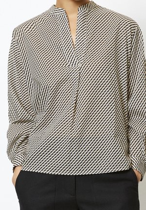 Vrouw draagt een beige en zwarte blouse met geometrisch patroon en lange mouwen en zwarte broek, handen in de zakken, staand tegen een witte achtergrond.