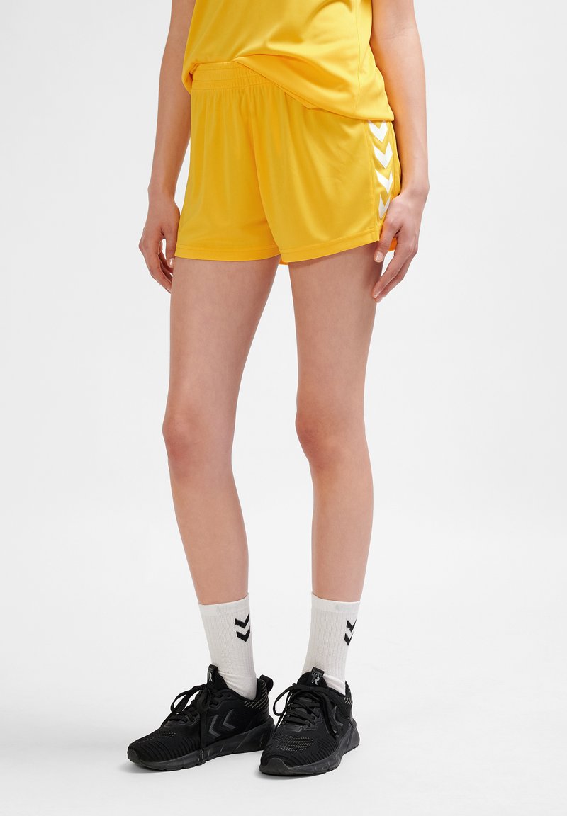 Gula sportshorts i lätt material med elastisk midja och vita chevrondetaljer på sidorna. Matchas med svarta skor och vita strumpor.