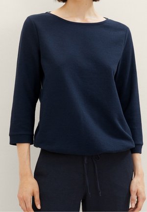 Vrouw draagt een donkerblauw gestructureerd shirt met lange mouwen en bijpassende broek met trekkoorden, gefotografeerd van schouders tot heupen tegen een neutrale achtergrond.