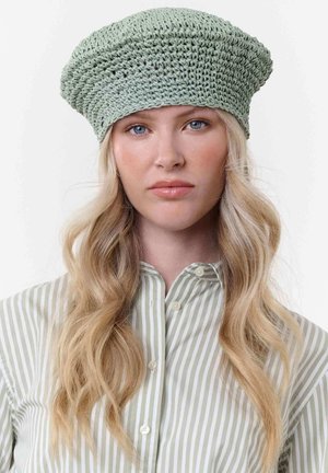 Young woman with long blonde hair wearing a light green crochet beret and a pale green striped button-up shirt, neutral expression.