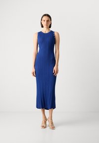 Robe midi bleue sans manches en maille côtelée avec des éléments décoratifs sur les épaules et une silhouette ajustée, accompagnée de talons beiges à lanières.