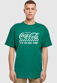 Žalias medvilninis marškinėlis su trumpomis rankovėmis, su baltu Coca-Cola logotipu ir šachmatų raštu, tekstas sako "Mėgaukitės Coca-Cola. Tai tikras dalykas."