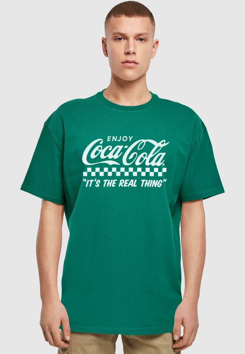 Žalias medvilninis marškinėlis su trumpomis rankovėmis, su baltu Coca-Cola logotipu ir šachmatų raštu, tekstas sako "Mėgaukitės Coca-Cola. Tai tikras dalykas."
