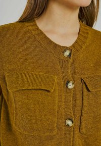 Cardigan giallo senape realizzato in tessuto a maglia testurizzato. Caratterizzato da un collo rotondo, due tasche anteriori a toppa e grandi bottoni circolari.