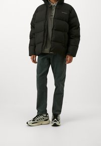 Person iført sort puffjakke, grå polo skjorte, mørkegrønne bukser og sort-hvide chunky sneakers står på hvid baggrund.