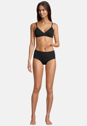 Frau steht barfuß und trägt ein schwarzes Bralette mit verstellbaren Trägern sowie schwarze, hoch taillierte Unterwäsche vor einem einfarbigen Hintergrund.