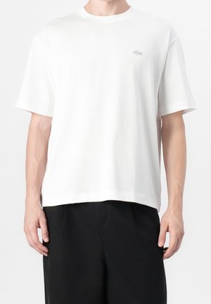Homme portant un t-shirt blanc uni à manches courtes avec un petit logo sur la poitrine gauche et un pantalon noir ample sur fond blanc.