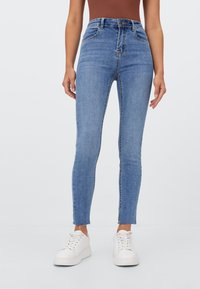 Jeans de denim azul ajustados con una cintura alta, que presentan un diseño clásico de cinco bolsillos y un dobladillo sin rematar en el tobillo, combinados con zapatillas blancas.