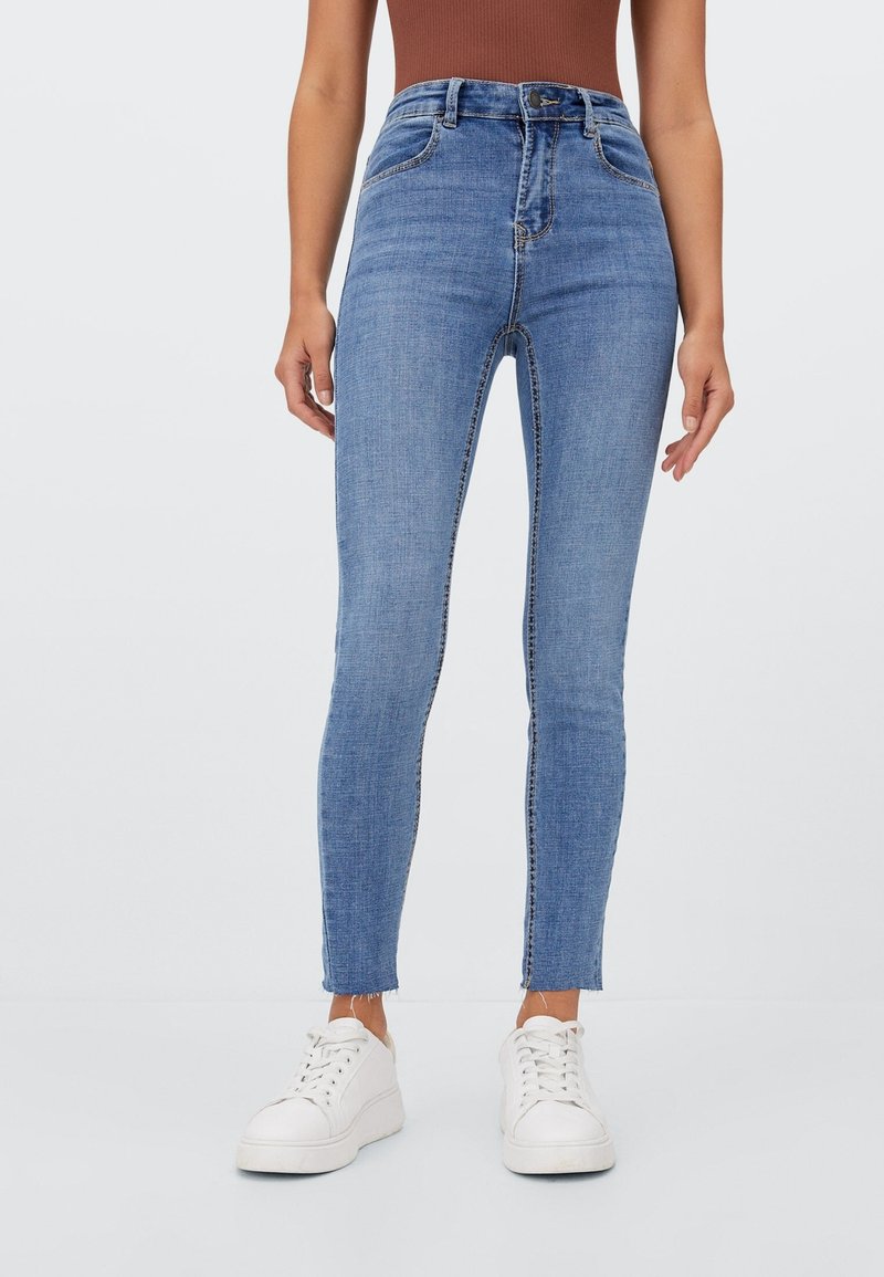 Jeans de denim azul ajustados con una cintura alta, que presentan un diseño clásico de cinco bolsillos y un dobladillo sin rematar en el tobillo, combinados con zapatillas blancas.