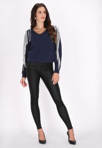 Marineblauer V-Ausschnitt-Pullover mit silberner Fransenverzierung an den Ärmeln, kombiniert mit schwarzen Leggings aus Kunstleder und schwarzen High-Heels.