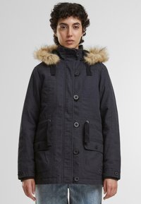 Črna parka jakna z kapuco obloženo z krznom, gumbnim zapiranjem, dvema sprednjima žepoma in nastavljivimi vrvicami. Ima teksturirano tkanino.