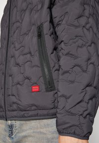 Gepolsterte dunkelgraue Jacke mit einer seitlichen Reißverschlusstasche, strukturiertem Stoff und einem roten Logoakzent am Saum. Lässiger Schnitt mit glatter Verarbeitung.