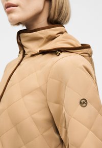 Κοντινό πλάνο ενός ατόμου που φοράει μπεζ καπιτονέ μπουφάν με καφέ περιθώρια και μικρό στρογγυλό μεταλλικό λογότυπο στον πάνω βραχίονα.