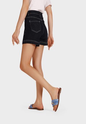 Shorts in denim scuro a vita alta con cuciture bianche, abbinati a sandali blu a infradito con un accento dorato. Vengono mostrate le gambe del modello.