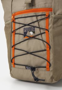 Khaki batoh s oranžovým zipem a akcenty, s černým designem bungee lana, síťovou boční kapsou a výrazným logem Puma v přední části.