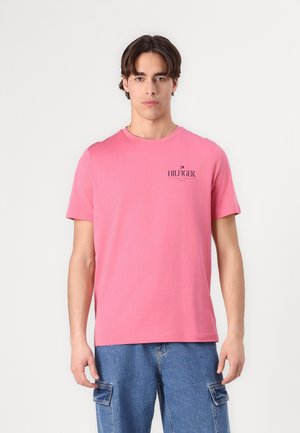 Rosa Baumwoll-T-Shirt mit kurzen Ärmeln, Rundhalsausschnitt und kleinem schwarzen "HILFIGER"-Logo auf der linken Brust. Das Modell trägt Jeansshorts.