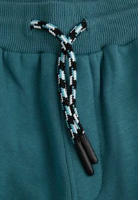 Turkusowy elastyczny pas z teksturowanym wykończeniem, wyposażony w plecioną czarną, białą i niebieską sznurówkę z plastikowymi końcówkami.