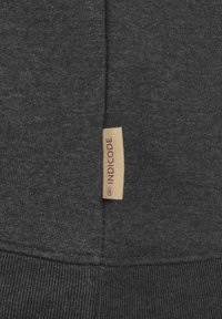 Grijze sweatshirt met een gladde textuur, geribde manchetten en een klein stoffen label met "INDICODE" in bordeauxrood op een beige achtergrond.