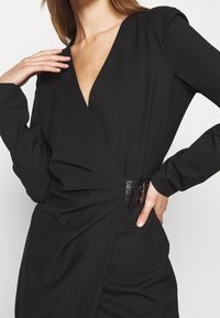 Schwarzes Kleid mit tiefem V-Ausschnitt, langen Ärmeln und gerafftem Bund. Verfügt über einen glänzenden schwarzen Gürtelakzent und eine glatte Textur.