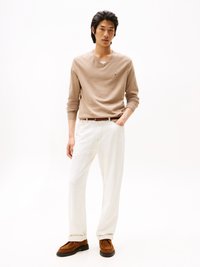 Pull en v beige associé à un pantalon blanc. Le mannequin porte des chaussures en suède marron et une ceinture marron. Design simple, texture lisse et couleurs douces.
